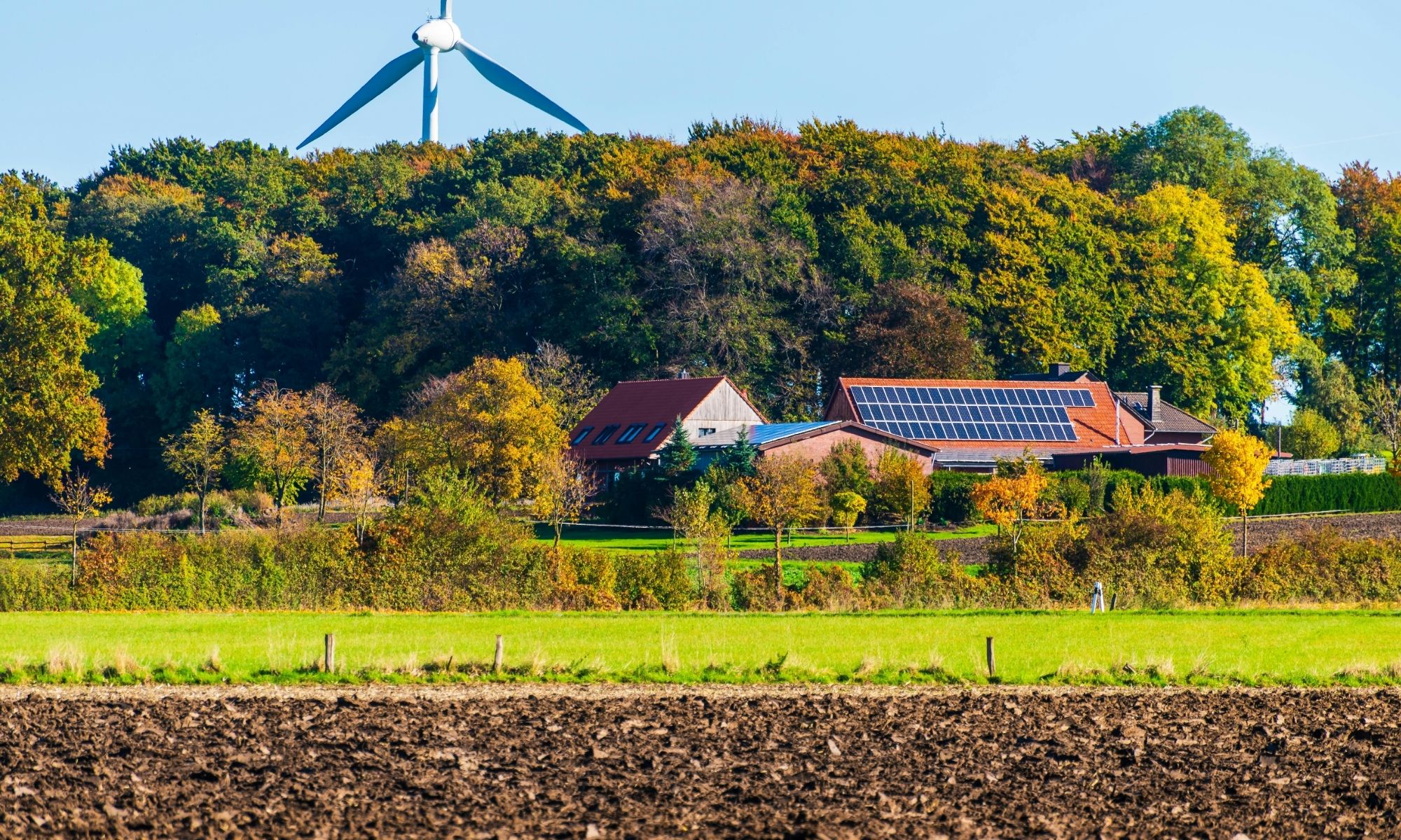 Rural house with solar panels near forested hill, wind turbine behind trees, and plowed soil—100 solutions for clean energy.