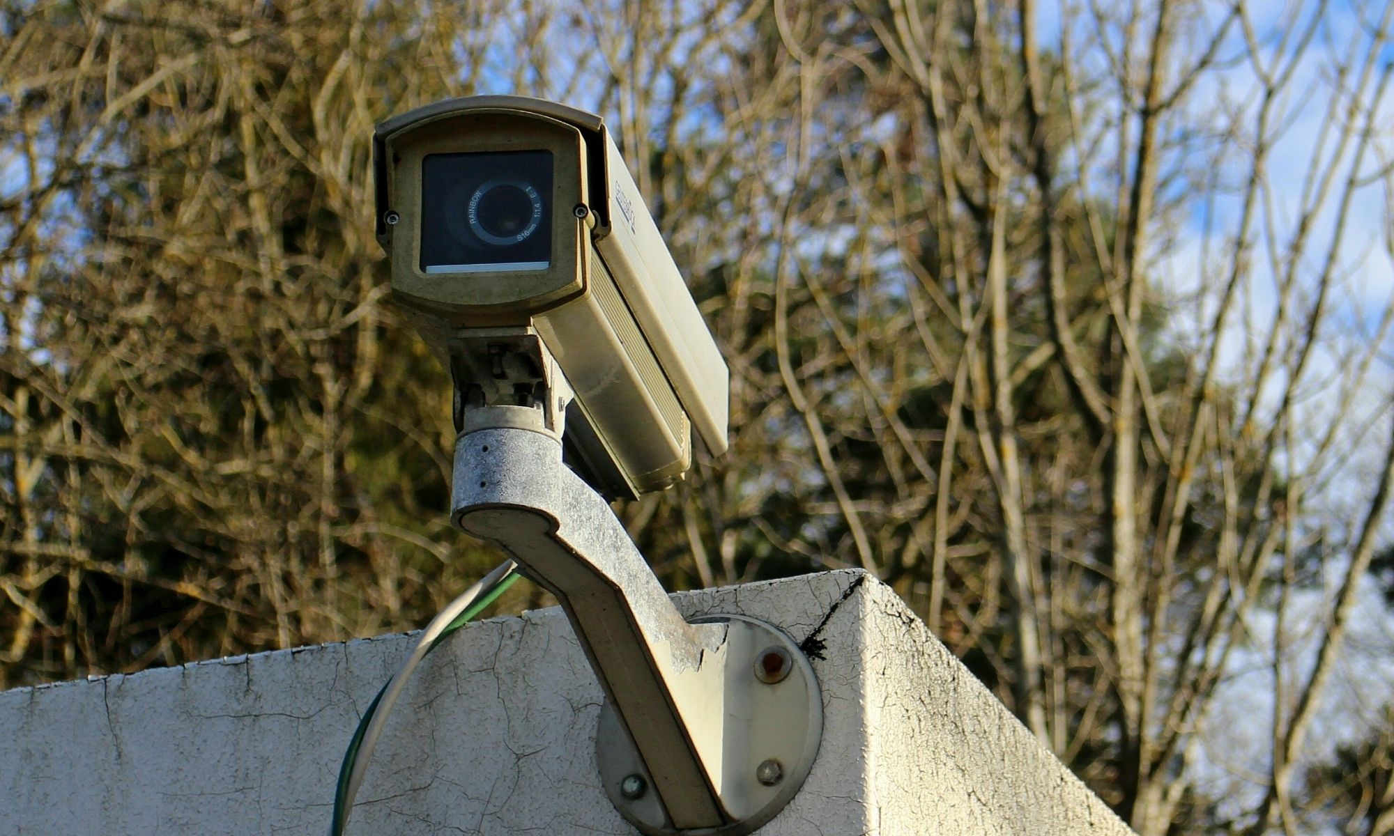 Security camera from 100 Solutions mounted on a concrete wall, angled up, with leafless trees and clouds in the background.