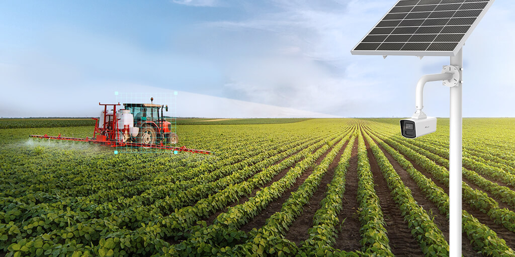 A tractor sprays crops as a solar-powered farm security camera is mounted on a pole in the foreground of a large field.