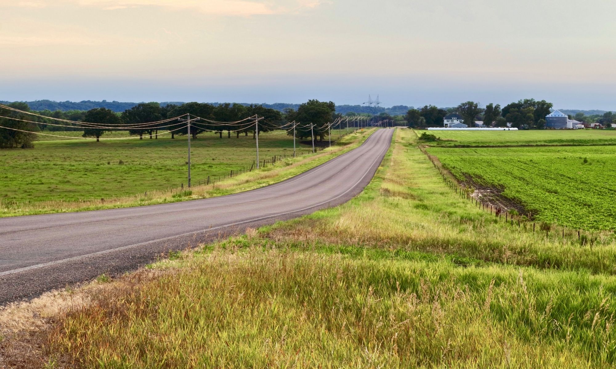 Two-lane rural road through green farmland, electric poles, distant trees, and buildings—100 Solutions for sustainable rural living.