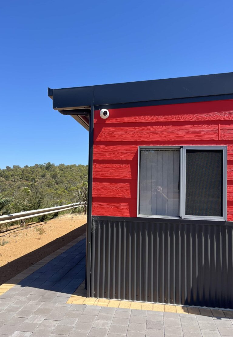 Red and gray building corner with security camera, window, railing, and paved walkway; business security is key.