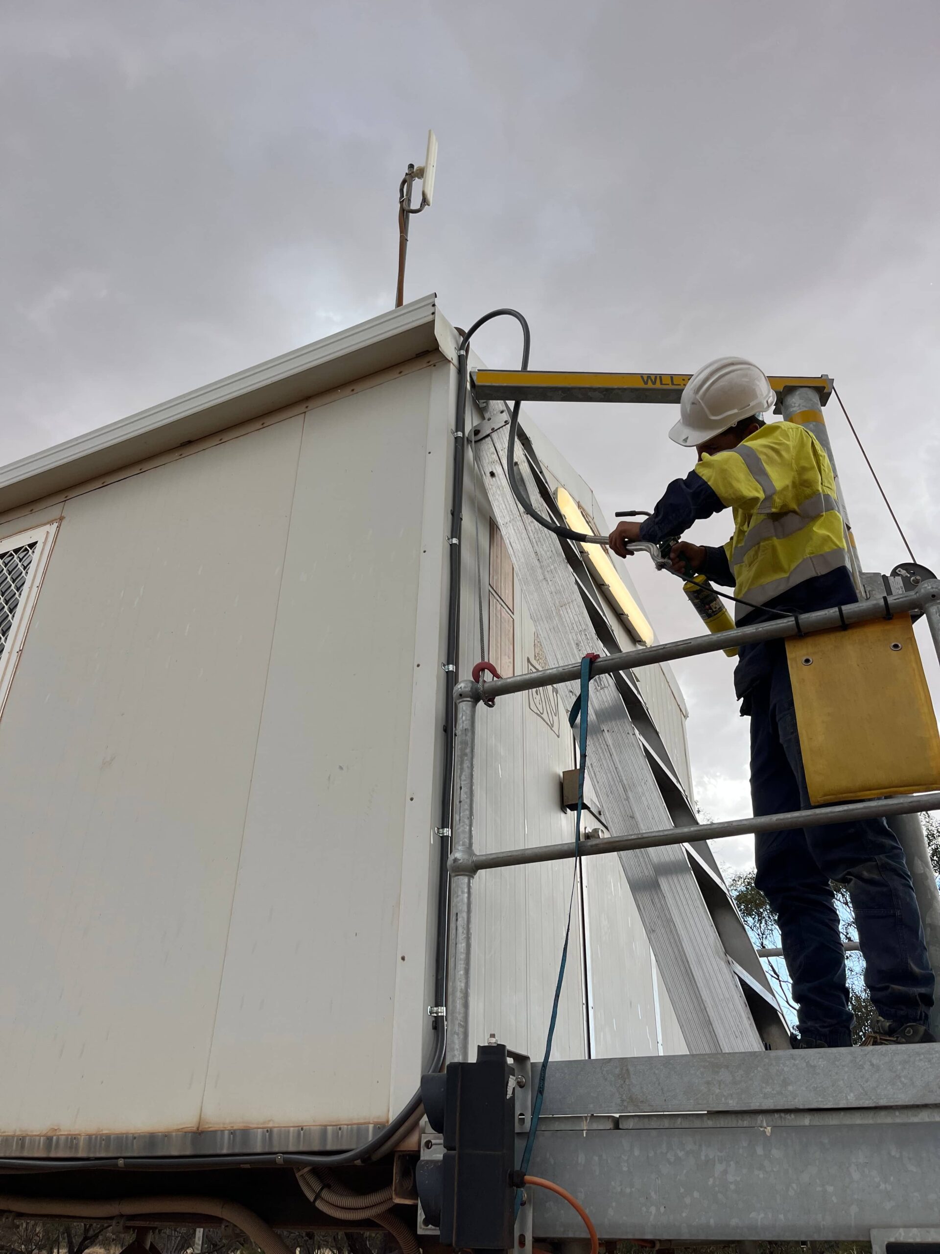 Worker in high-visibility jacket using power tool on commercial building yellow lift for security.