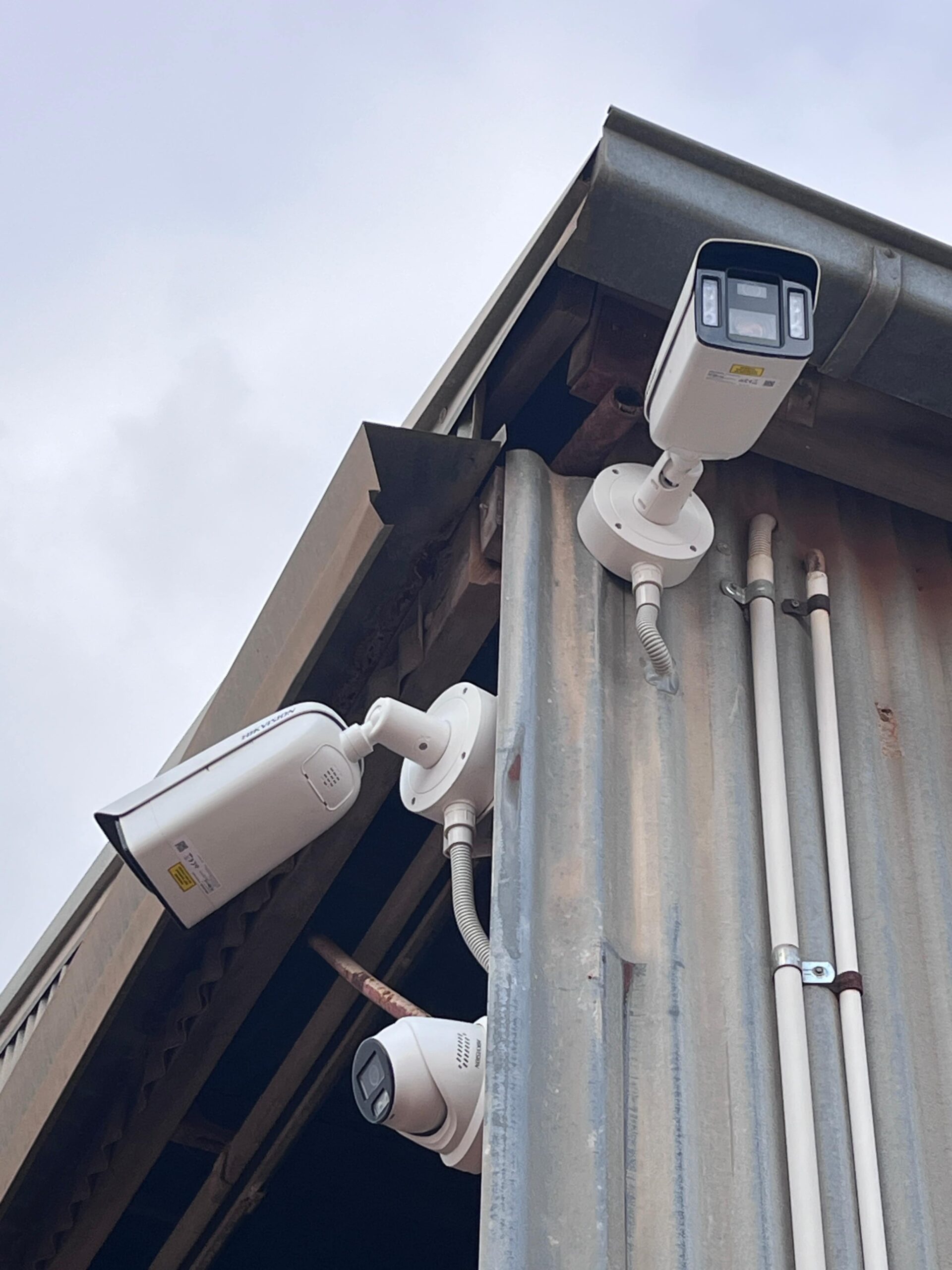 Three security cameras on a corrugated metal building provide commercial security under a cloudy sky.