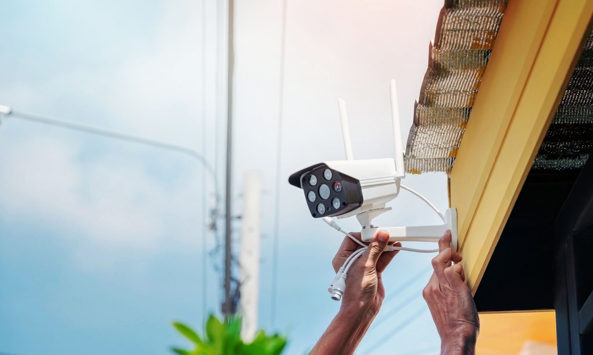 Installing a security camera under a yellow building’s roof edge to enhance commercial security; power lines visible.