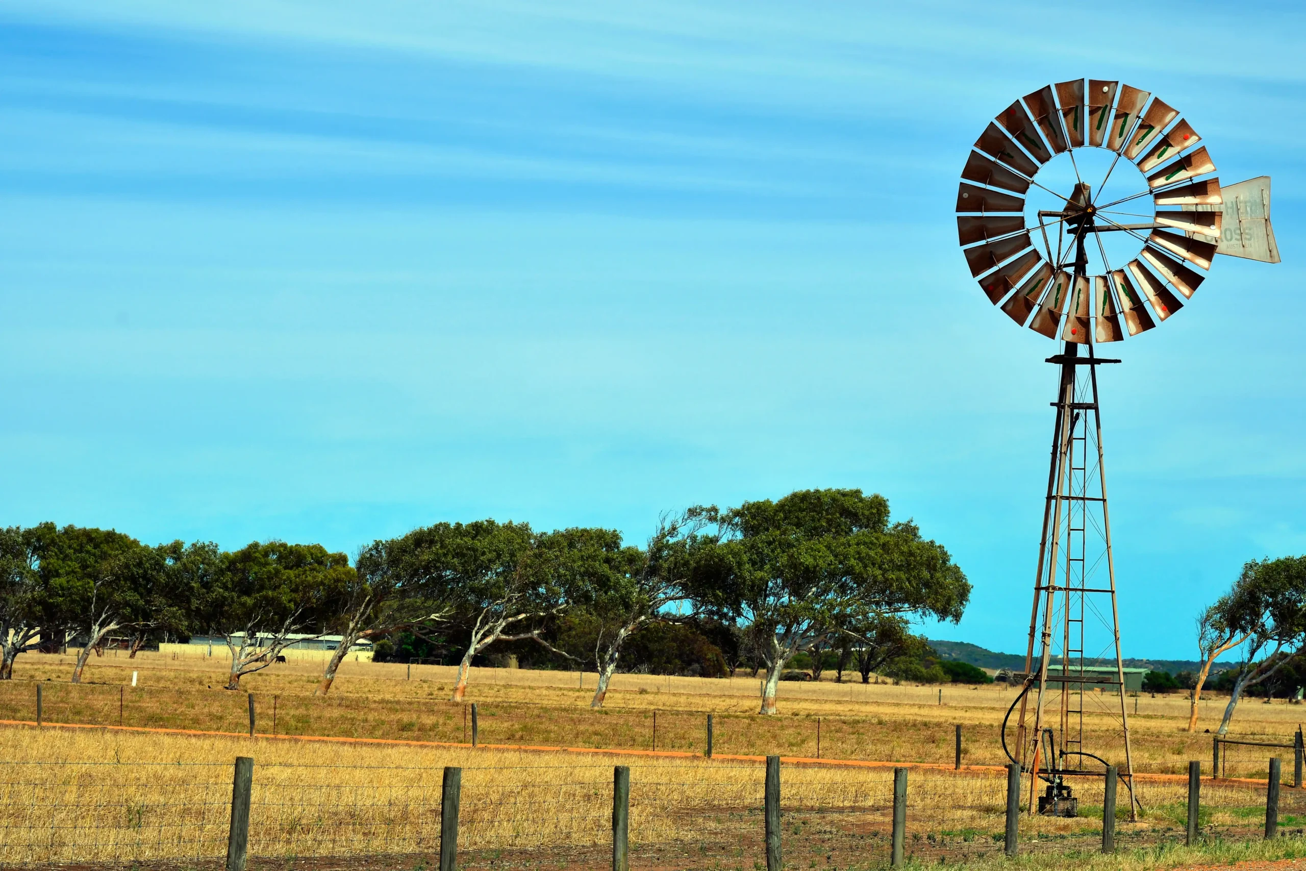 Metal windmill in dry, grassy field with fence, trees, blue sky near remote fertilizer manufacturing facility.