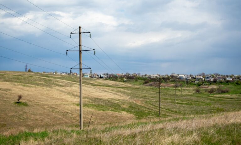 Wooden utility poles with power lines stretch across a grassy field, village in the distance, cloudy sky.